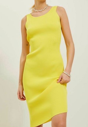 Mujer con vestido amarillo midi sin mangas y acanalado, sujetando el dobladillo con una mano, lleva un collar de cadena y una pulsera sobre fondo liso.