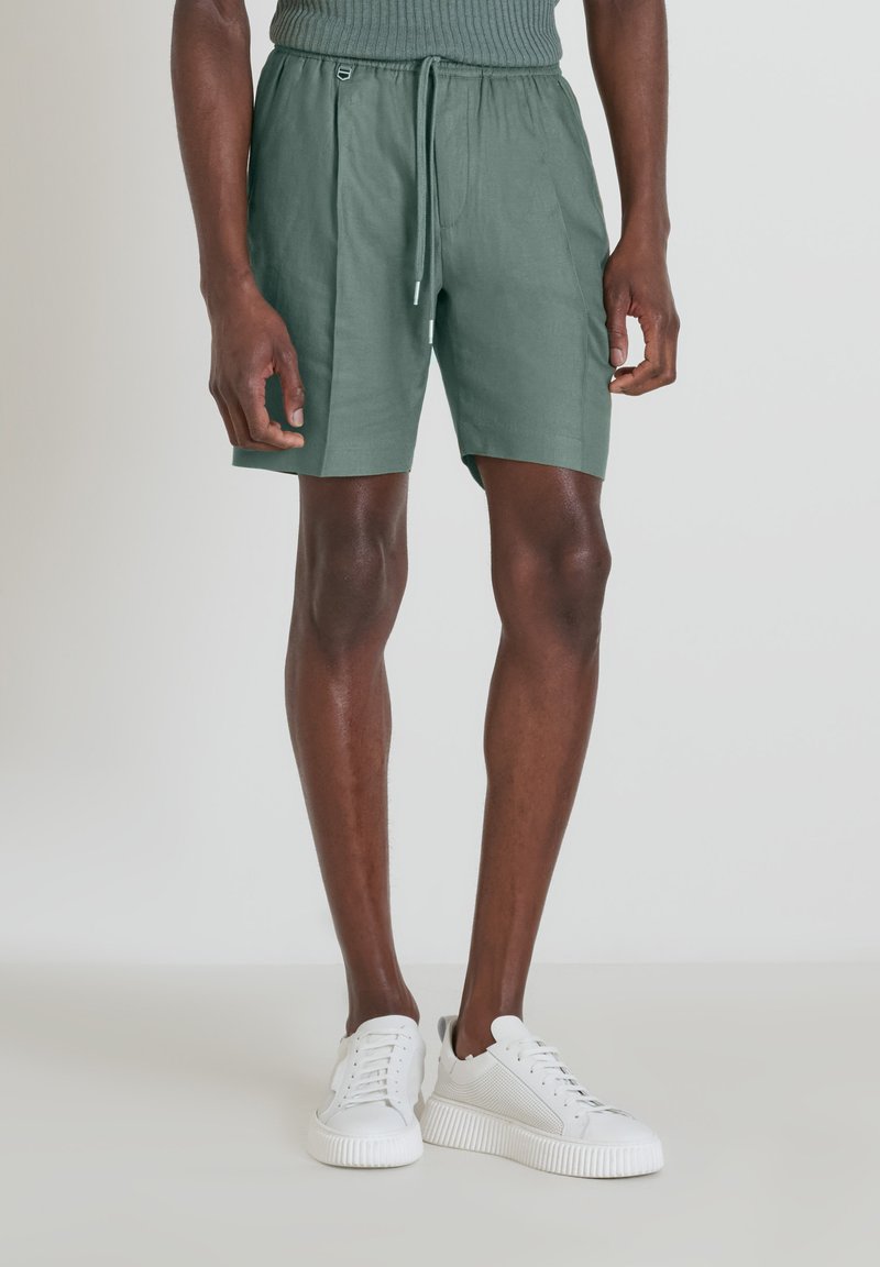 Pantaloni corti in lino verde con vita elastica, chiusura con lacci e tasche laterali, abbinati a sneakers bianche. Texture liscia.
