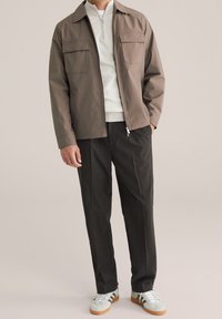 Uomo che indossa una giacca zip-up color taupe, un maglione bianco a mezze cerniera, pantaloni grigio scuro e sneakers bianche con suole color beige e strisce marroni.