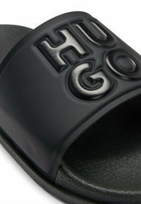 Black slide sandal with a textured footbed and a wide strap featuring embossed "HUGO" lettering. Made from synthetic materials; matte finish.