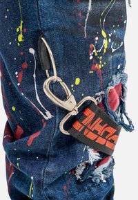 Pantalon en denim bleu avec un design de peinture éclaboussée en rouge, jaune et blanc. Comprend un fermoir en métal argenté et une sangle noire avec des lettres rouges.
