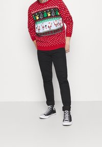 Roter Pullover mit weißen Punkten, versehen mit einem festlichen Muster aus Bäumen, Schneemännern und dem Weihnachtsmann. Kombiniert mit schwarzen Hosen und schwarzen High-Top-Sneakern.