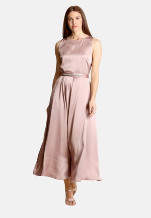 Donna che indossa un abito senza maniche in raso rosa blush lungo fino al pavimento, con cintura gioiello e tacchi aperti, in piedi davanti a uno sfondo semplice.