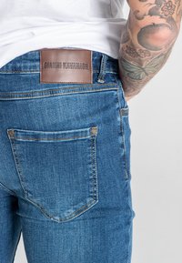 Vaqueros de denim azul con corte slim. Presentan un parche de cuero marrón en la cintura grabado con "GIANNI KAVANAGH" y bolsillos traseros estándar.