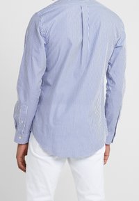 Pasiasta koszula z długim rękawem w kolorze niebieskim i białym, o prostym kroju, z plisą z tyłu i guzikami przy mankietach, noszona na białych spodniach.
