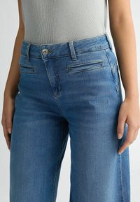 Jean en denim bleu avec une coupe droite, présentant deux poches avant, une fermeture à bouton et des détails de couture subtils. Texture lisse.