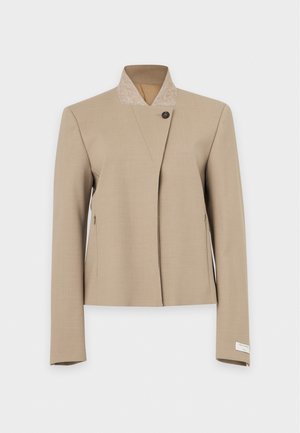 Un mannequin aux longs cheveux foncés porte une veste beige cintrée et un pantalon à jambes larges, ainsi que des chaussures à talons marron. Il se tient devant un fond clair uni.