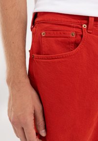 Røde denimjeans med klassisk fem-lomme-design, som har gullfarget hardware og et teksturert, slitesterkt stoff. Nærbilde av side-lommen.