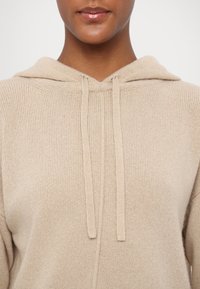Gros plan sur une personne portant un sweat à capuche beige en tricot avec cordons, montrant le cou, les lèvres et la partie supérieure de la poitrine.