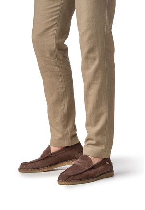 Mocassini in pelle scamosciata marrone con design cucito, suola piatta e tacco leggero. Abbinati a pantaloni beige chiaro con taglio stretto.