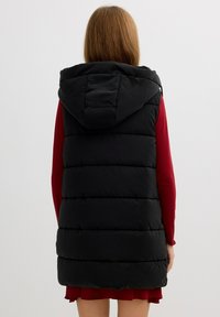 Chaleco de plumas negro con capucha, que presenta secciones acolchadas y una textura suave. Usado sobre una prenda de manga larga roja.