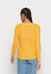 Pull en tricot de couleur jaune vif, avec des manches longues et un col large, finition texturée, associé à un jean bleu.
