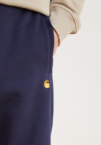 Navyblaue Sweatpants aus weichem Material mit einem goldenen, gestickten Logo am linken Oberschenkel und einer Seitentasche.