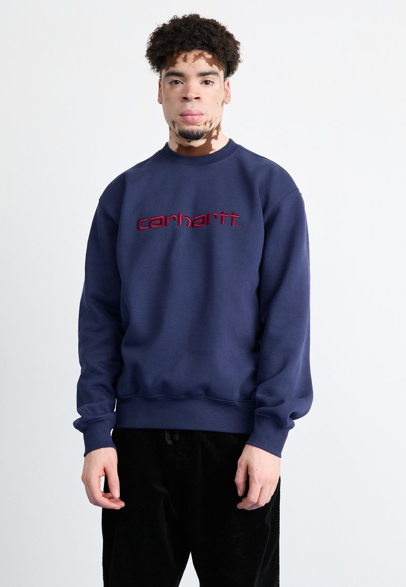 Sudadera azul marino con puños y dobladillo de canalé, con un destacado logo bordado en rojo "carhartt" en el pecho.