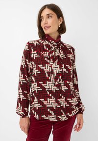 Long-sleeve blouse in a red, black, and white zigzag pattern. Features a bow tie collar and soft fabric with a smooth texture.