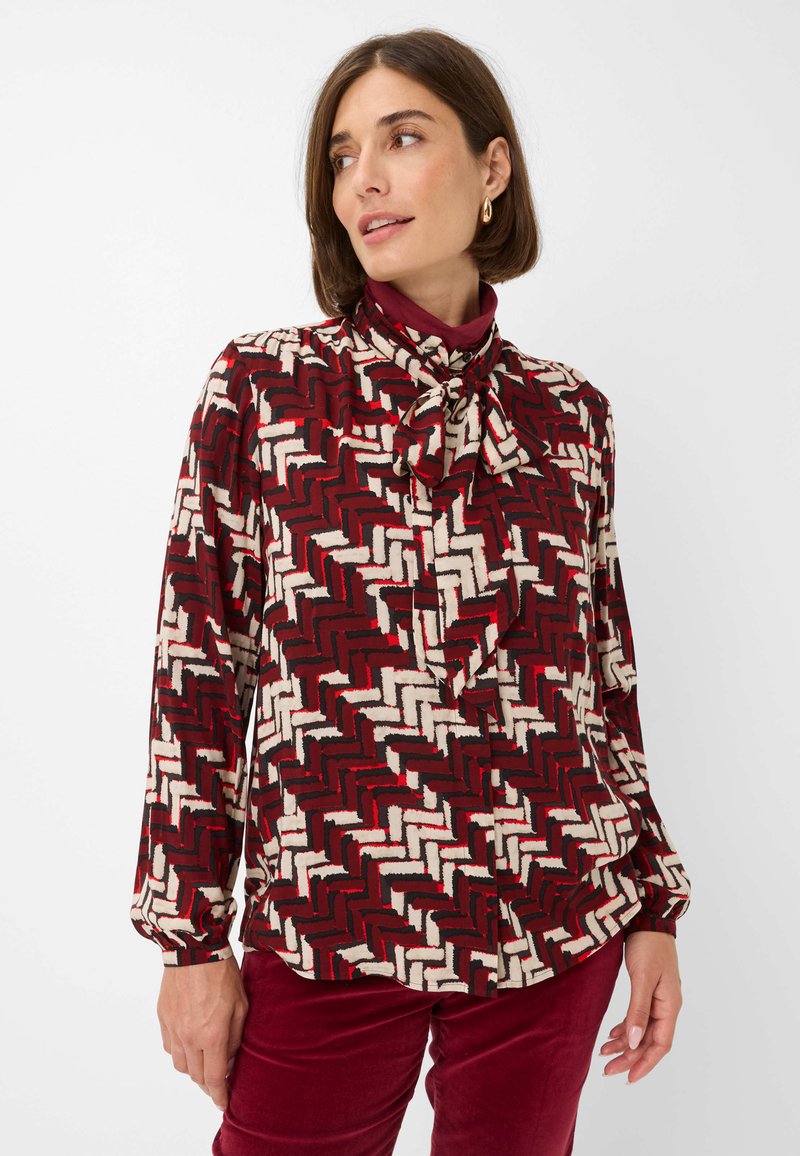 Long-sleeve blouse in a red, black, and white zigzag pattern. Features a bow tie collar and soft fabric with a smooth texture.