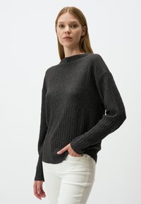 Pull en tricot gris foncé avec un motif côtelé structuré, un col haut et des manches longues, associé à un pantalon blanc.