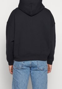 Personne portant un sweat à capuche noir uni et un jean en denim bleu clair vue de dos sur un fond neutre.