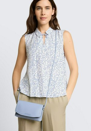 Vrouw draagt mouwloze witte blouse met blauwe bloemprint, beige broek en een lichtblauw schoudertas, staand met handen in de zakken.