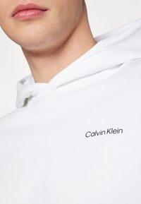 Felpa bianca con tessuto texturizzato, cappuccio con cordino e logo ricamato "Calvin Klein" in nero sul petto.