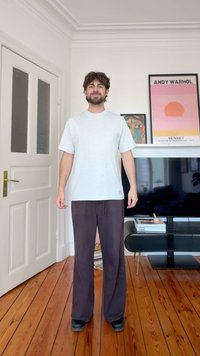 Νέος άνδρας με καστανά μαλλιά και μούσια, φορώντας ανοιχτό γκρι μπλουζάκι και σκούρο παντελόνι, στέκεται σε ξύλινο πάτωμα στο σαλόνι με έργα τέχνης και τηλεόραση στο βάθος.