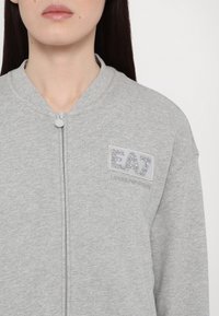 Femme portant une veste zippée gris clair avec un logo EA7 Emporio Armani scintillant sur le côté gauche de la poitrine.