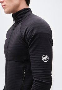 Νεαρό άτομο που φοράει μαύρο μπουφάν με φερμουάρ και ψηλό γιακά, με λευκό λογότυπο μαμούθ στο πάνω μέρος του μανικιού, μπροστά σε απλό φόντο.