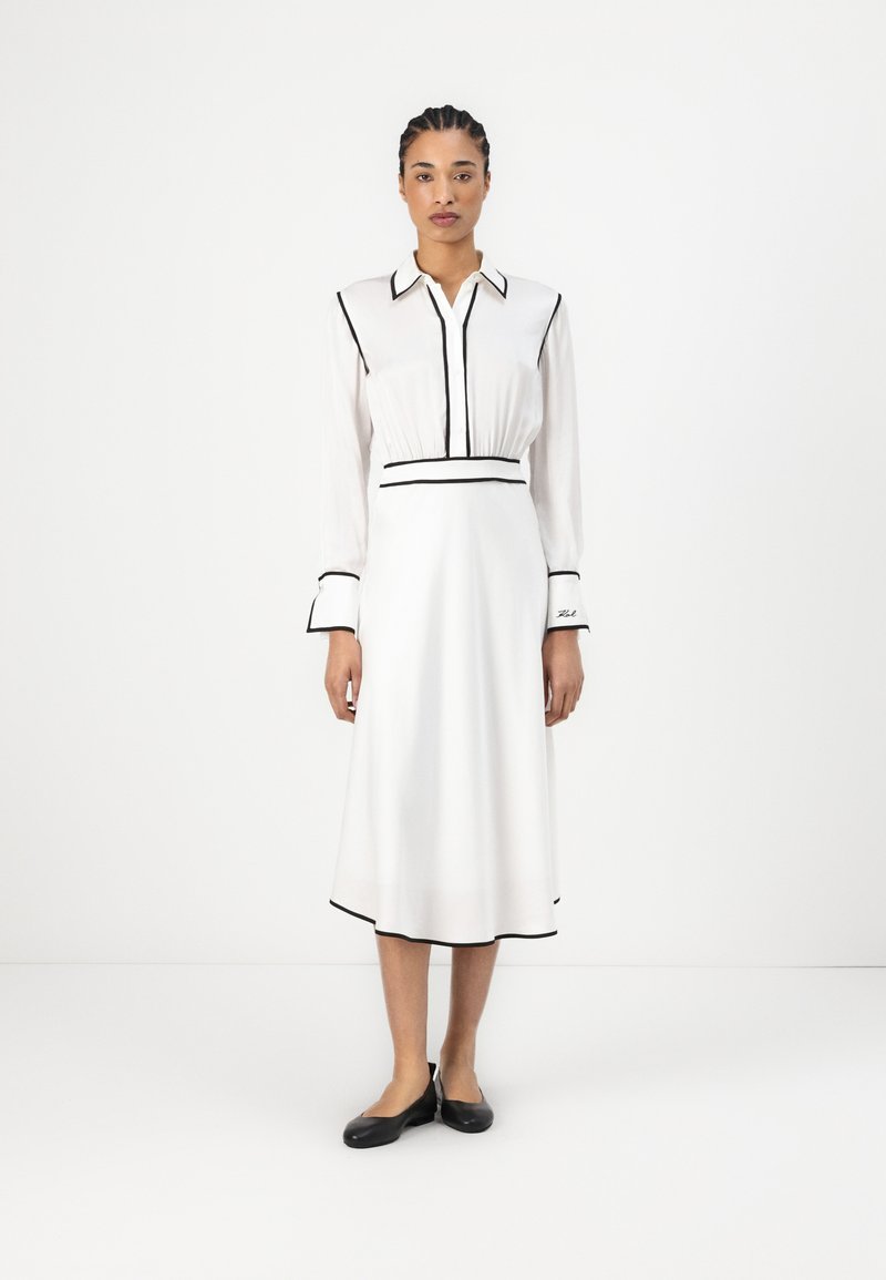 Robe blanche avec des accents noirs, comportant un col, un devant à boutons, des manches longues et une taille ajustée. Jupe évasée en A.