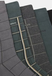 Satz von vier Paar Socken: grau, grün, dunkelgrün und schwarz. Merkmale sind kontrastierende Muster und strukturierte Bänder nahe den Oberteilen.