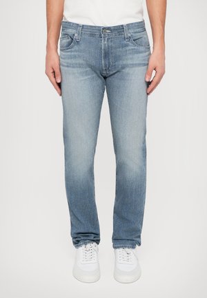 Personne portant un jean droit bleu clair et des baskets blanches, debout, les mains détendues le long du corps devant un fond uni.