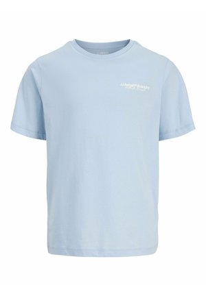 Garçon blond debout, une main dans la poche, portant un t-shirt bleu clair, un jean bleu large et des baskets beiges sur fond blanc.