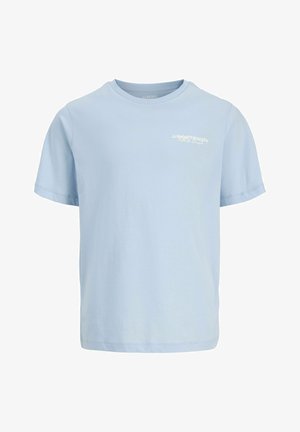 Blond jongetje staat met één hand in zijn zak, draagt een lichtblauw t-shirt, losse blauwe spijkerbroek en beige sneakers op een witte achtergrond.