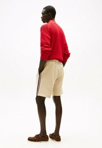 Maglione rosso a trama, pantaloni corti beige con righe laterali blu navy e rosse, e mocassini marroni. Modello visto da dietro, con le mani in tasca.