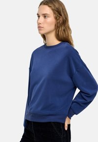 Niebieski bawełniany sweter z okrągłym dekoltem, opadającymi ramionami i prążkowanymi mankietami. Gładka tekstura i luźny krój, zestawiony z ciemnymi spodniami.