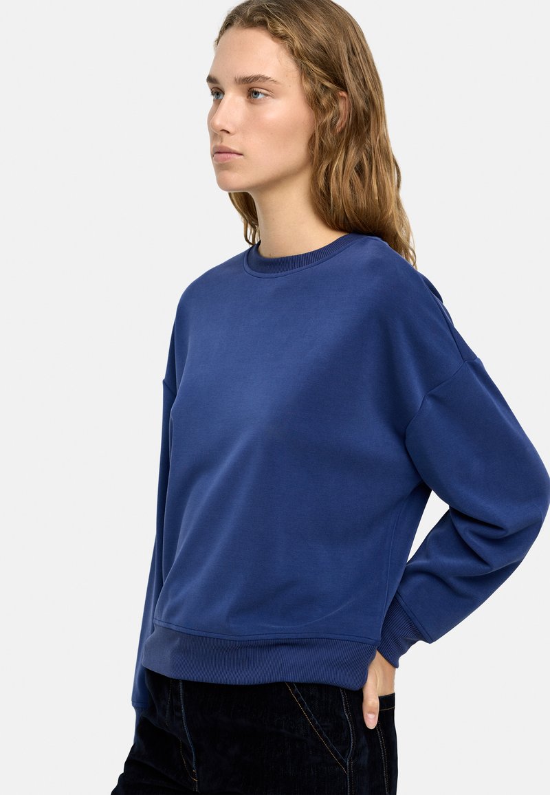 Niebieski bawełniany sweter z okrągłym dekoltem, opadającymi ramionami i prążkowanymi mankietami. Gładka tekstura i luźny krój, zestawiony z ciemnymi spodniami.