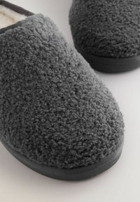 Un paio di pantofole pelose grigio scuro con tessuto simile alla lana e suole in gomma nera su una superficie chiara.