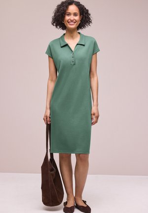 Uśmiechnięta kobieta o kręconych włosach, ubrana w zieloną sukienkę do kolan zapinaną na guziki, trzyma brązową zamszową torbę i ciemne płaskie buty.