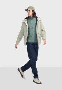 Hellgrüne Kapuzenjacke über einem dunkelgrünen Fleecemantel mit Reißverschluss, kombiniert mit dunkelblauen Jeans und beigen Sneakers. Das Modell trägt eine passende Mütze.