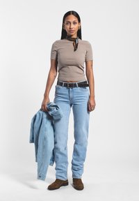 Top ajustado beige, jeans azul claro, sosteniendo una chaqueta de mezclilla. Cinturón negro, botas altas marrones. Tela suave, corte relajado, estilo casual.