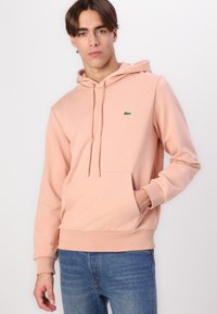 Sudadera rosa claro con cordones ajustables, bolsillo frontal, puños acanalados y un pequeño logo verde. Combinada con vaqueros azules.