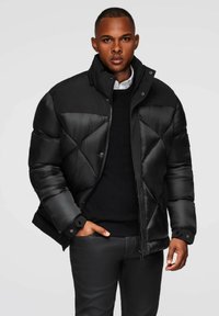 Giacca a vento nera con un design geometrico trapuntato, colletto alto e chiusure a pressione. Indossata sopra un maglione nero a maglia e pantaloni scuri.