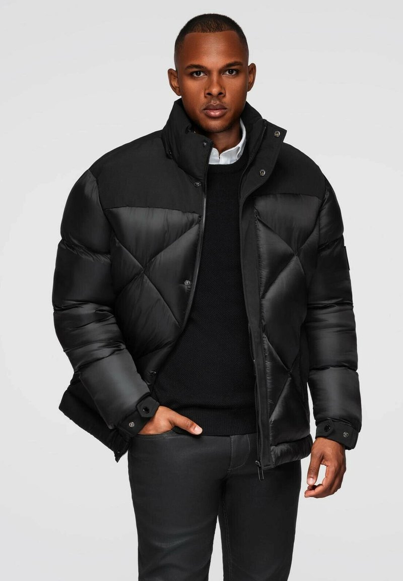 Giacca a vento nera con un design geometrico trapuntato, colletto alto e chiusure a pressione. Indossata sopra un maglione nero a maglia e pantaloni scuri.