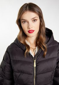 Giacca a vento nera con cappuccio, design trapuntato, zip dorata, indossata sopra una camicia bianca. I capelli sono mossi e le labbra sono truccate di rosso.