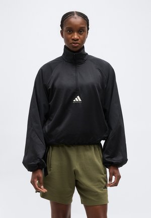 Νεαρό άτομο με πλεγμένα μαλλιά, φορώντας μαύρο μακρυμάνικο πουλόβερ και σορτς χακί, στέκεται μπροστά σε απλό ανοιχτόχρωμο φόντο.