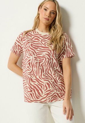 Kobieta o długich blond włosach, ubrana w beżowo-rdzawą koszulę w paski zebry z krótkim rękawem oraz jasne spodnie, stojąca na tle jednolitej ściany.