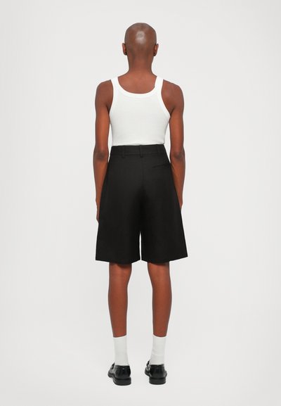 Svart shorts med skreddersydd design, laget av et glatt stoff, med en baklomme, brukt med en hvit tank top og sokker.