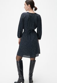 Kleid in Navy mit floralem Muster, langen Ärmeln und gebundenem Taillenband. Der Stoff ist leicht, kombiniert mit knielangen schwarzen Stiefeln.