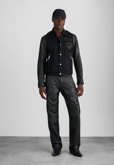 Jachetă din suede negru și piele, cu două buzunare cu fermoar și accente cu logo, asortată cu pantaloni cargo din piele neagră. O șapcă elegantă completează aspectul.