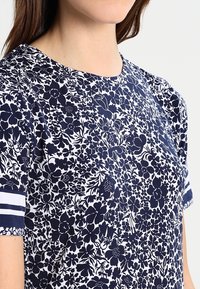 Haut à motifs floraux bleu marine et blanc avec des manches courtes, présentant des rayures blanches horizontales aux bords des manches et une texture de tissu lisse.