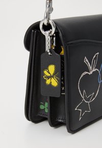Sac en cuir noir avec des motifs brodés, incluant un papillon jaune, une pomme et des étoiles. Doté d'une chaîne métallique et d'un détail d'étiquette.